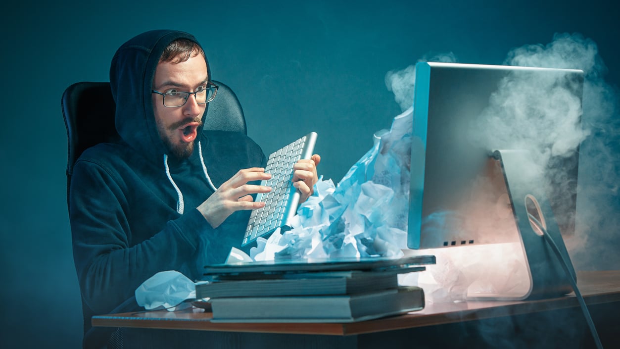 Man by a computer shocked holding a keyboard, crumbled papers and smoke coming out of the computer 